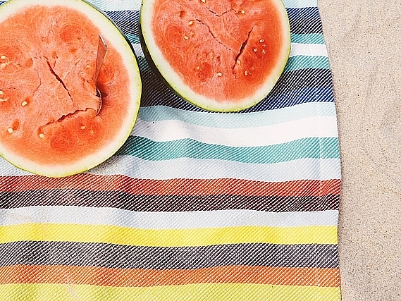 Zwei Hälften einer Wassermelone liegen auf einem bunt gestreiften Tuch am Strand.