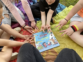 Mehrere Kinder sitzen auf einer bunten Picknickdecke bei Jumpers Döbeln und strecken ihre Hände in die Mitte zu einem Kinderbuch mit dem Titel „Ich bin fast genau wie du“, das auf der Decke liegt.