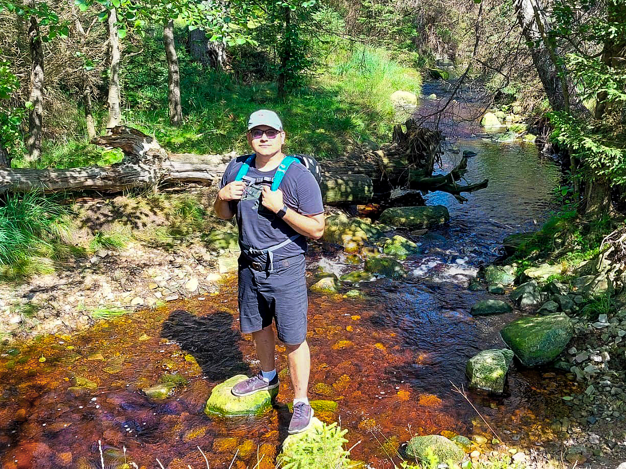 Rick Gensch steht mit Rucksack und heller Kappe auf einem Stein in einem flachen Bach im Wald, umgeben von grünen Bäumen, Gras und einem umgestürzten Baumstamm.
