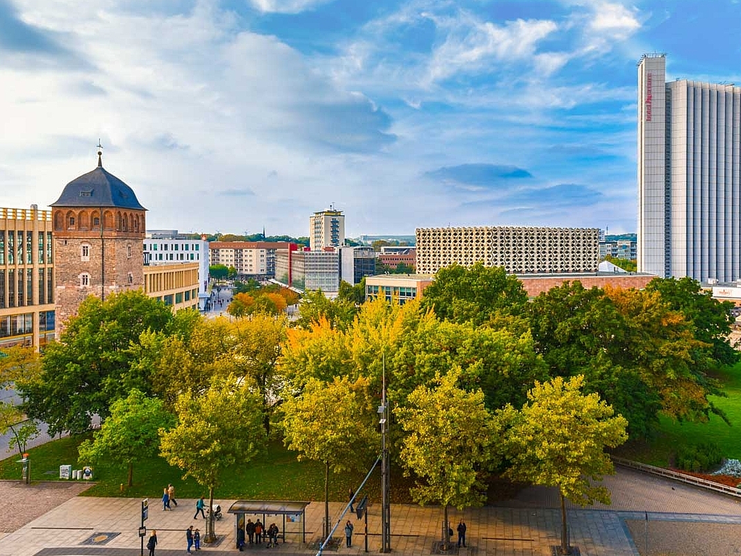 Grünfläche mit Bäumen und im Hintergrund mit hellblauem Himmel die Innenstadt von Chemnitz mit dem roten Turm