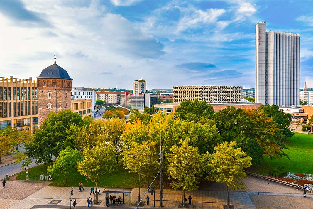 Grünfläche mit Bäumen und im Hintergrund mit hellblauem Himmel die Innenstadt von Chemnitz mit dem roten Turm