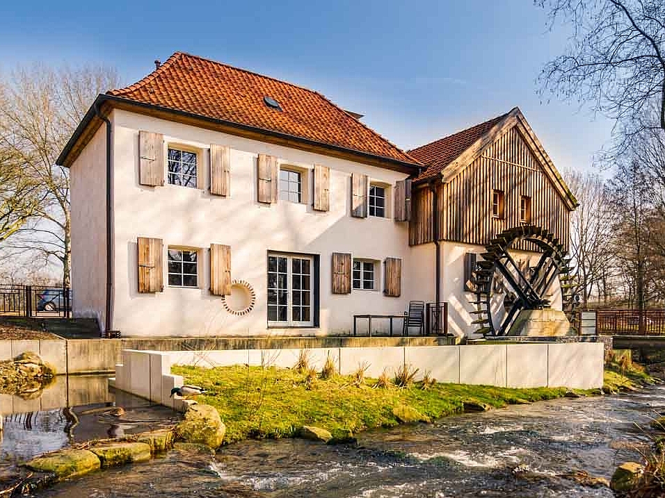 Cremefarbenes Haus mit Mühlenrad in Moers und davor ist ein kleiner Fluss zu sehen