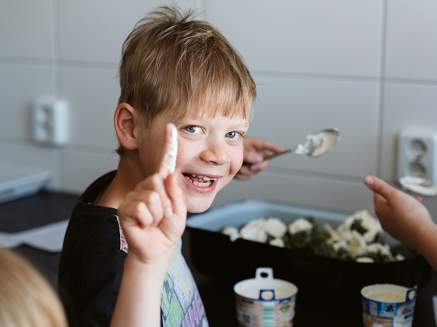 Ein lachendes Kind steht in der Küche bei Jumpers Döbeln, hält einen Finger mit weißer Creme in die Kamera und blickt fröhlich nach vorne. Im Hintergrund sind eine Auflaufform mit Zutaten sowie weitere Kinderhände mit Löffeln zu sehen.