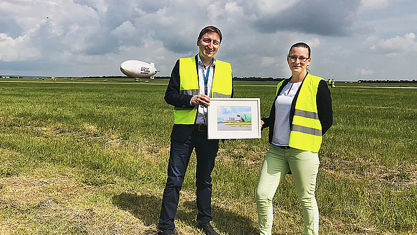TAG Wohnen Standortleiter Claudius Oleszak und Teamleiterin Yvonne Beyer auf dem Erfurter Flughafen mit Luftschiff „Theo“ im Hintergrund: Das Wohnungsunternehmen unterstützt die Stadtmarketing-Aktion mit dem Slogan „Wohnen in Zukunft“.