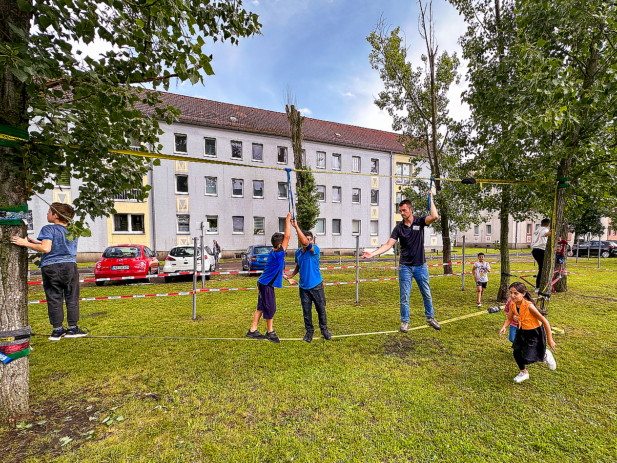 Kinder balancieren mit Christian Mende auf einer Slackline im grünen Innenhof eines Wohnquartiers, umgeben von Bäumen und Mehrfamilienhäusern, bei einem Bewegungsangebot der Jumpers im TAG-Viertel.