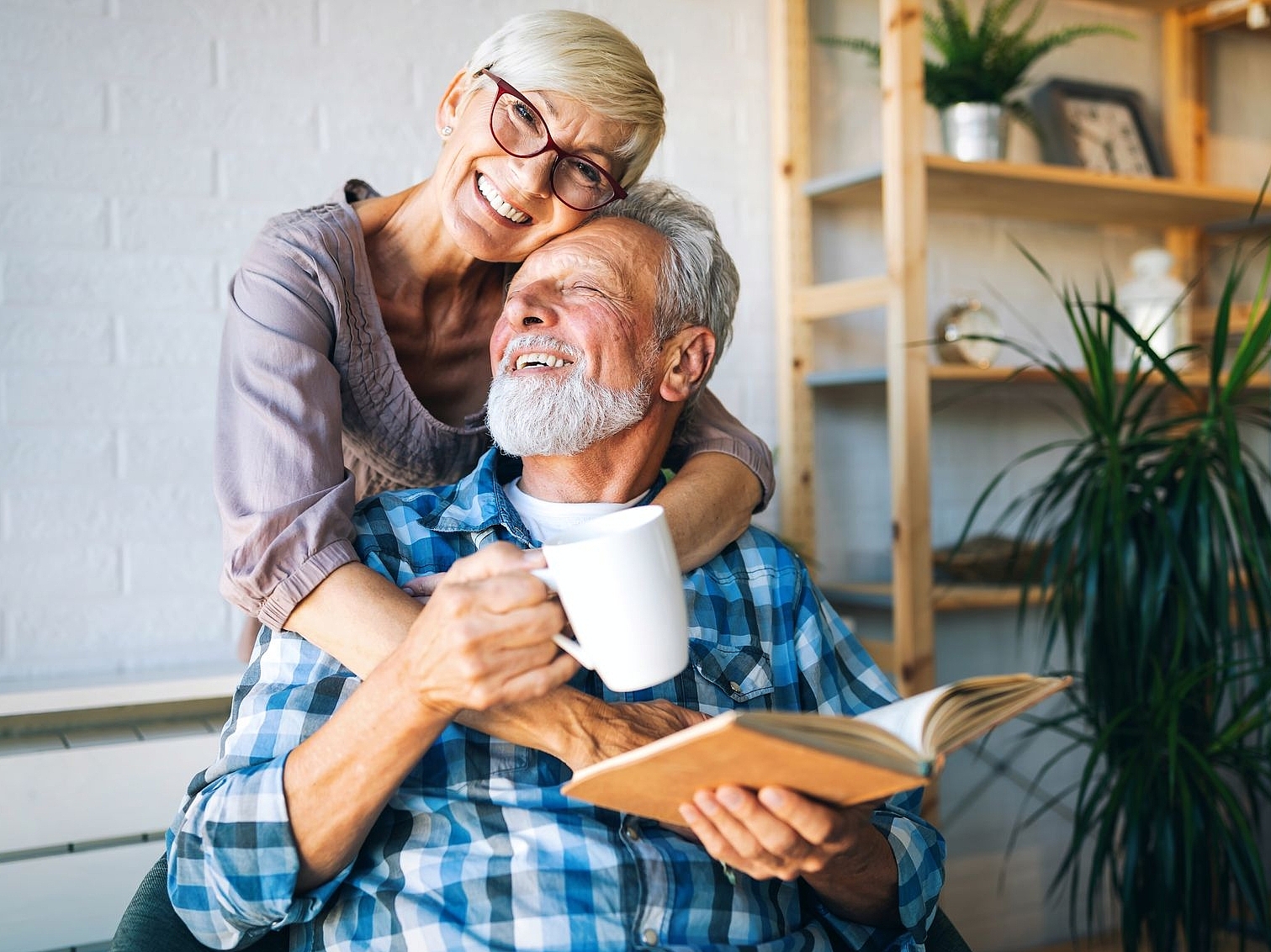 Eine ältere Frau die einen älteren Mann lachend und liebevoll von hinten umarmt.