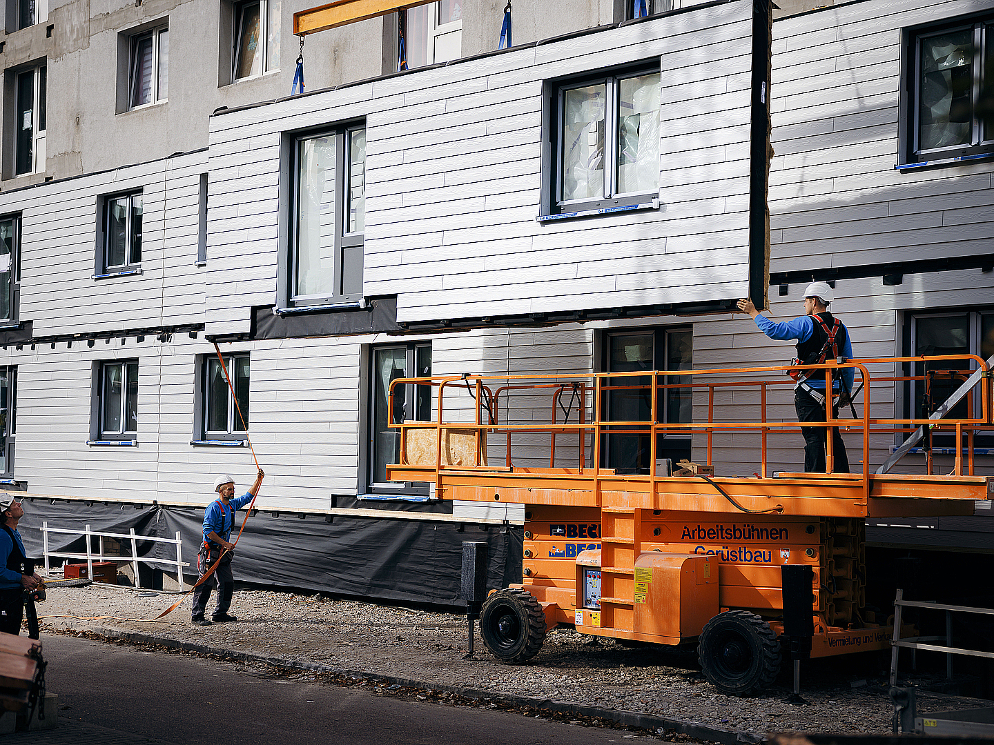 Auf der Baustelle geht es jetzt auf die Hebebühne und ein Fachmann montiert die Fassadenmodule auf der zweite Etage.