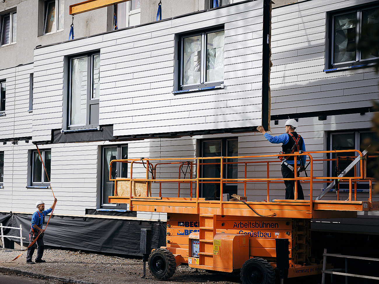 Auf der Baustelle geht es jetzt auf die Hebebühne und ein Fachmann montiert die Fassadenmodule auf der zweite Etage.