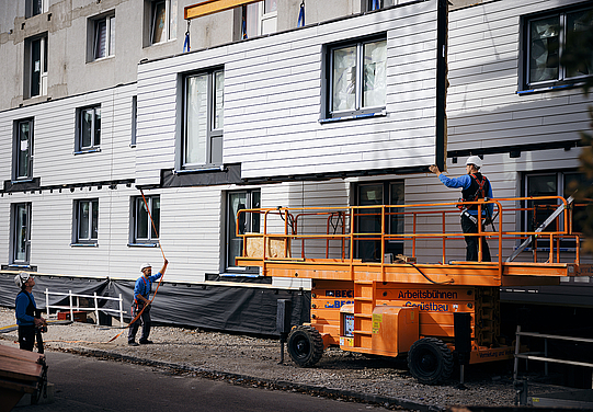 Auf der Baustelle geht es jetzt auf die Hebebühne und ein Fachmann montiert die Fassadenmodule auf der zweite Etage.