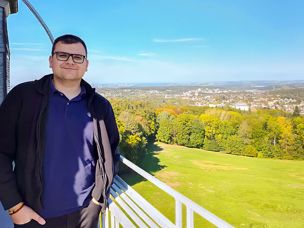 Rick Gensch steht auf einer Aussichtsplattform und blickt in die Kamera. Im Hintergrund sieht man eine weitläufige Landschaft mit einer Stadt im Hintergrund. einer Stadt in der Ferne unter blauem Himmel.