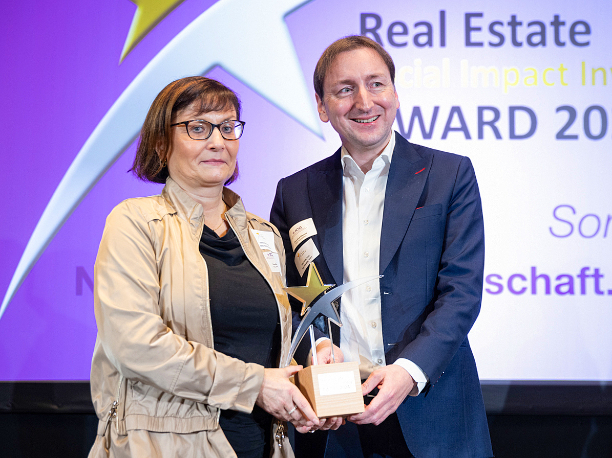 Claudia Hoyer, TAG Vorstand und Claudius Oleszak, Leiter Business & Change Development stehen auf der Bühne und halten gemeinsamden den Real Estate Social Impact Investing Award 2024 (SII 2024) fest 