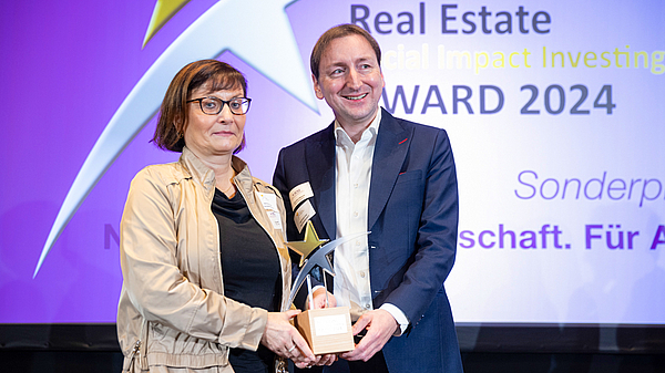 Claudia Hoyer, TAG Vorstand und Claudius Oleszak, Leiter Business & Change Development stehen auf der Bühne und halten gemeinsamden den Real Estate Social Impact Investing Award 2024 (SII 2024) fest