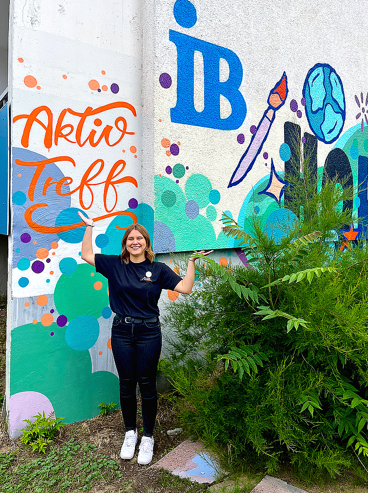 Das Bild zeigt Jolina Seide vor einer Hauswand, die mit Graffiti besprüht ist. Das Graffiti enthält das Logo des IB.