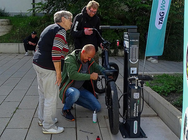 Ein interessierter Passant repariert ein Fahrrad an der neuen Fahrrad-Reparaturstation, während eine ältere Passantin und ein weiterer Besucher die Funktionen am Berliner Platz neugierig studieren und die Reparatur beobachten.