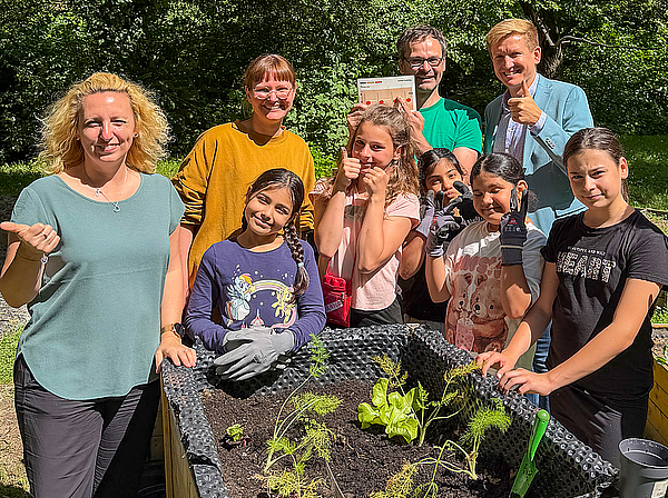 Bei Sonnenschein stehen der Regionalleiter Thüringen der TAG Wohnen Eric Frey, Ackercoach Dirk Fehse, die Teamleitung Immobilienbewirtschaftung in Gera, Dominique Nündel, sowie Jumpers gemeinsam um ein frisch bepflanztes Hochbeet im Gemeinschaftsgarten und präsentieren stolz die jungen Gemüsepflanzen.