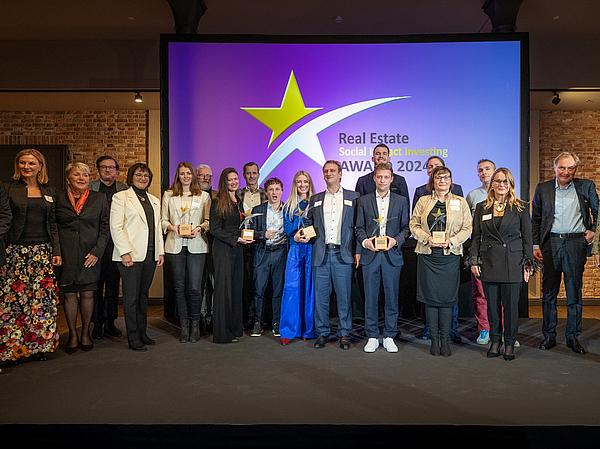 Gewinnerinnen und Gewinner stehen gemeinsam mit den fünf Preisträgerinnen und Preisträgern des Real Estate Social Impact Investing Award 2024 nach der Preisvergabe auf eine großen Bühne.