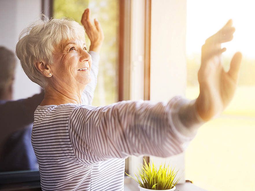 Eine Frau steht mit weit ausgebreiteten Armen vor einem geöffneten Fenster.