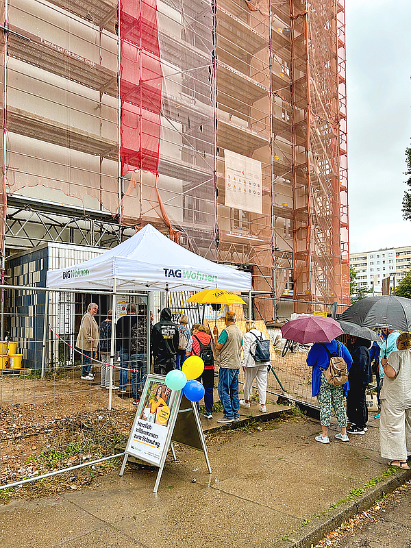 Vor dem eingerüsteten Zehngeschosser in der Otto-Nagel-Straße warten Besucherinnen und Besucher mit Regenschirmen am TAG-Wohnen-Zelt, um sich beim Schautag über die modernisierten Wohnungen zu informieren.