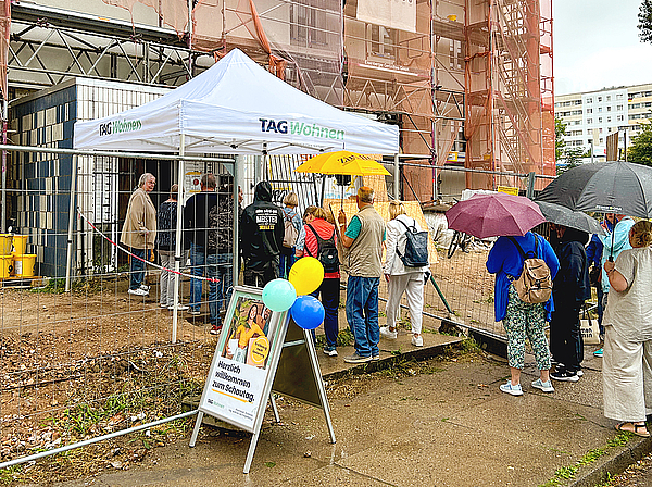 Vor dem eingerüsteten Zehngeschosser in der Otto-Nagel-Straße warten Besucherinnen und Besucher mit Regenschirmen am TAG-Wohnen-Zelt, um sich beim Schautag über die modernisierten Wohnungen zu informieren.