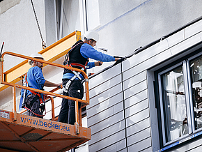 Zwei Handwerker stehen auf einer erhöhten Plattform und montieren das neue Fassadenelement an das Gebäude