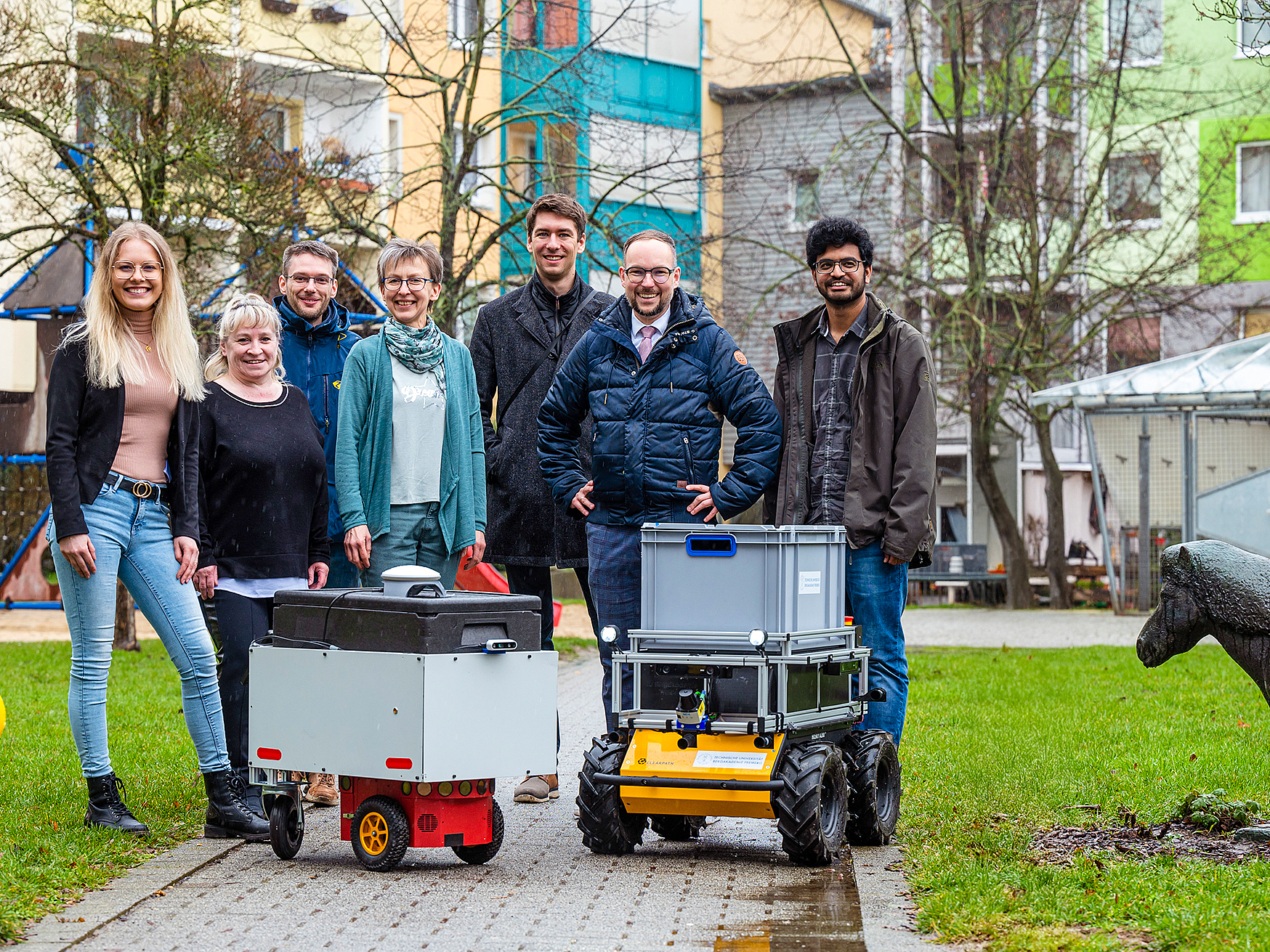 Eine Menschengruppe steht hinter dem neuen Lieferroboter, im Bildhintergrund ist ein Plattenbau zu sehen.  