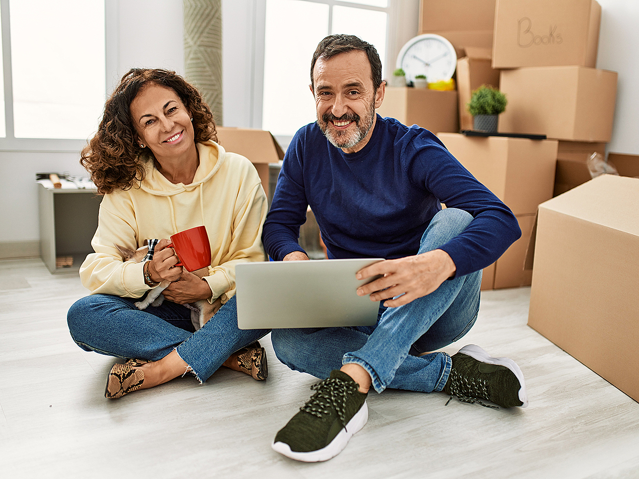 Ein glückliches Paar zieht in eine neue Wohnung ein, sie lächeln in die Kamera und im Hintergrund sind viele Umzugskartons