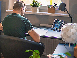Ein junger Mann sitzt in einer Wohnung an einem Schreibtisch und arbeitet.