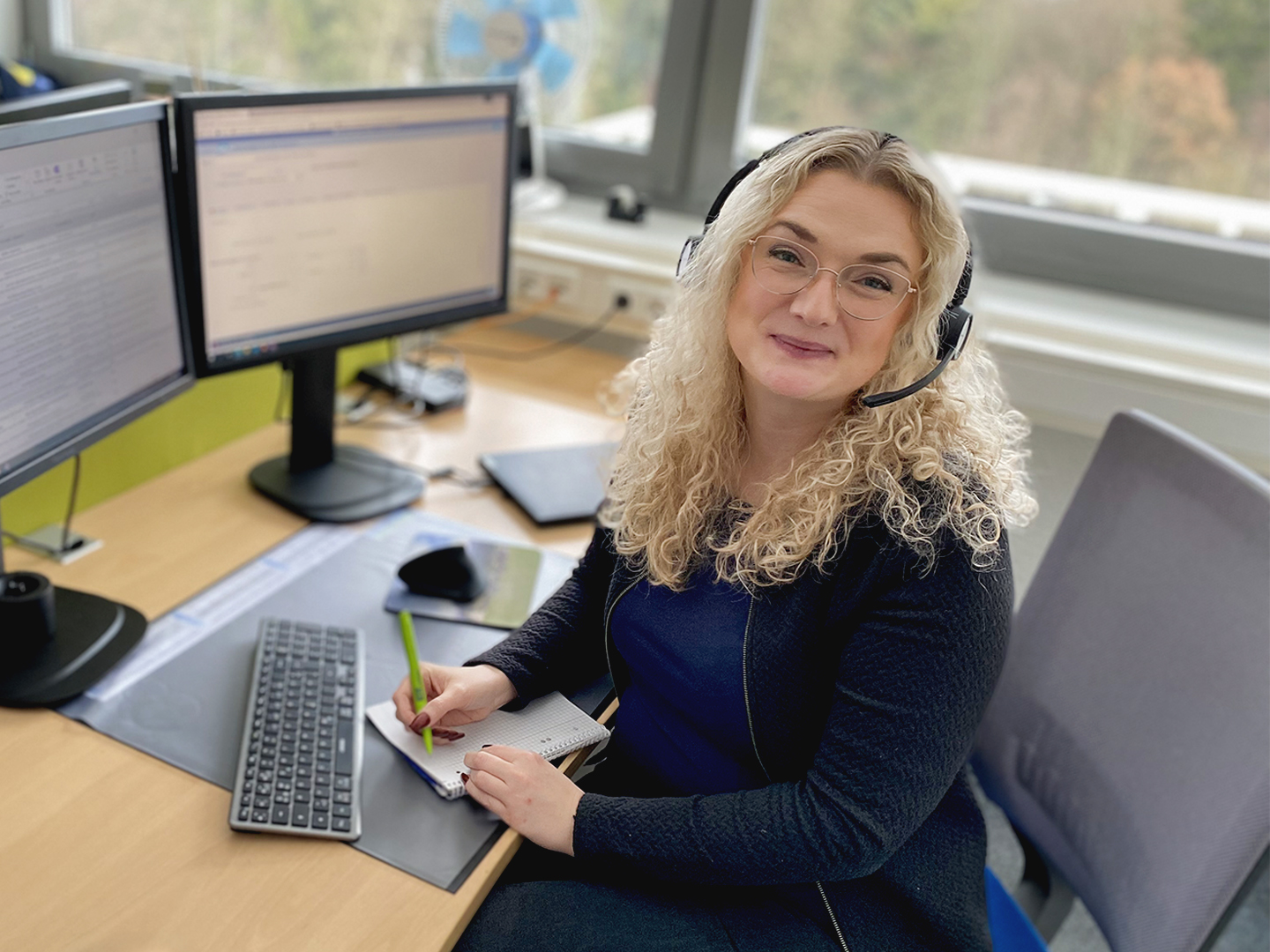 Das Bild zeigt Dagmar Schmidt am Schreibtisch sitzend vor zwei Monitoren und mit Headset auf dem Kopf