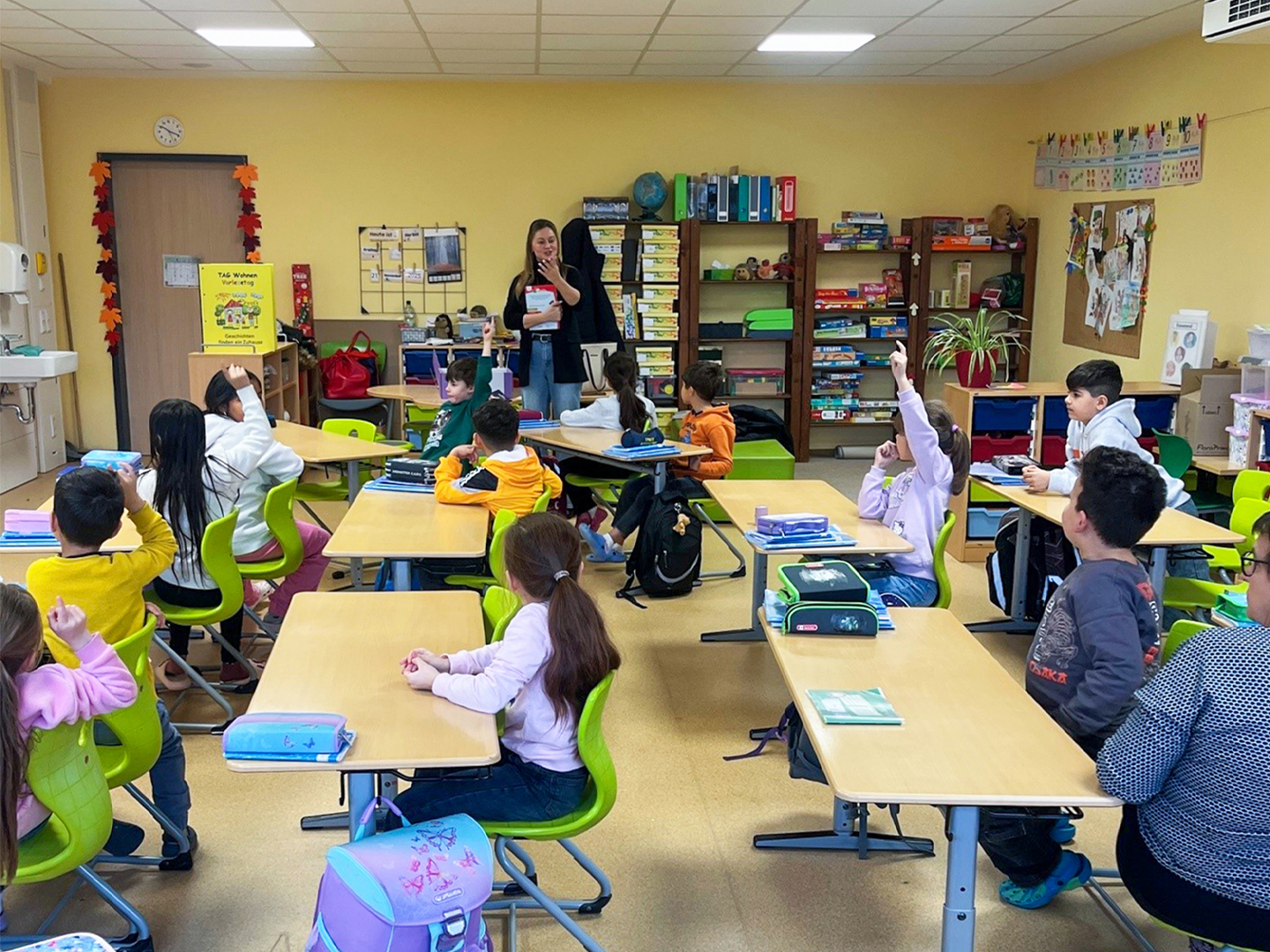 Das Bild zeigt ein Klassenzimmer. Die Kinder schauen nach vorne zu einer Frau, die ein Buch in der Hand hält. Einige Kinder melden sich.